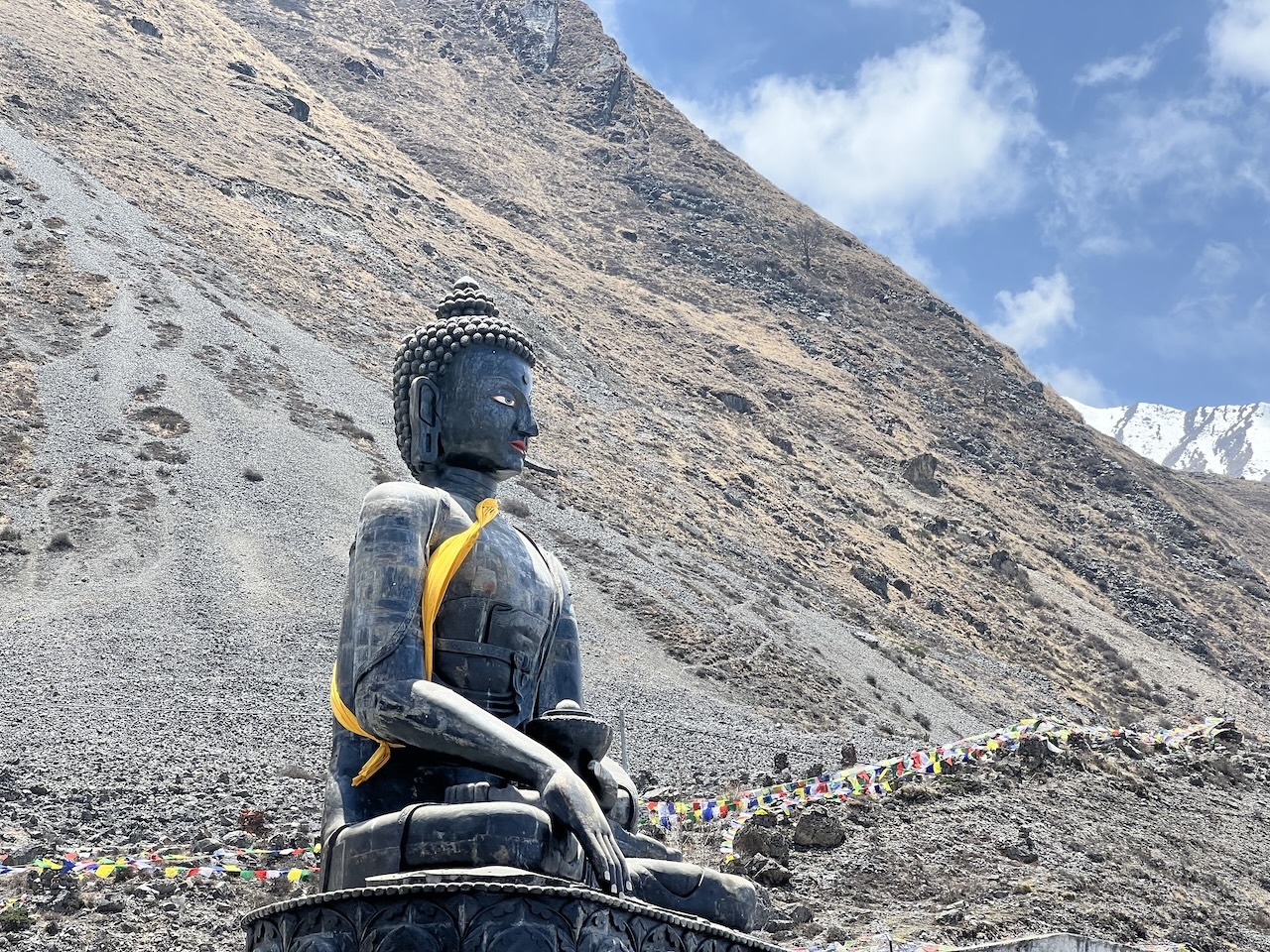 Monumento di Buddha a Muktinah Statua del Buddha seduto con drappo giallo, bandiere di preghiera e pendii rocciosi sullo sfondo