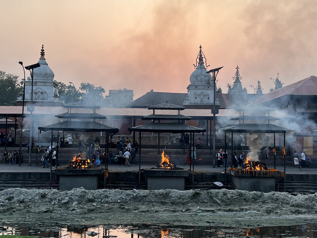 Pashupatinath Temple di Kathmandu Roghi funerari su bracieri lungo il fiume, fumo denso e padiglioni con templi bianchi sullo sfondo.