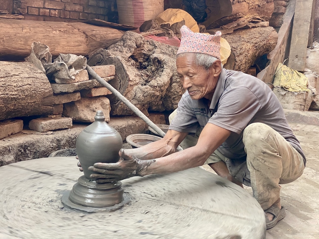 Vasaio a Thimi Anziano vasaio al tornio che modella un vaso d'argilla con mani coperte di fango, cappello e sfondo laborioso.