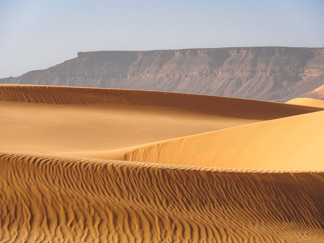 In primo piano dune di sabbia ondulate con increspature, sullo sfondo un altopiano roccioso 