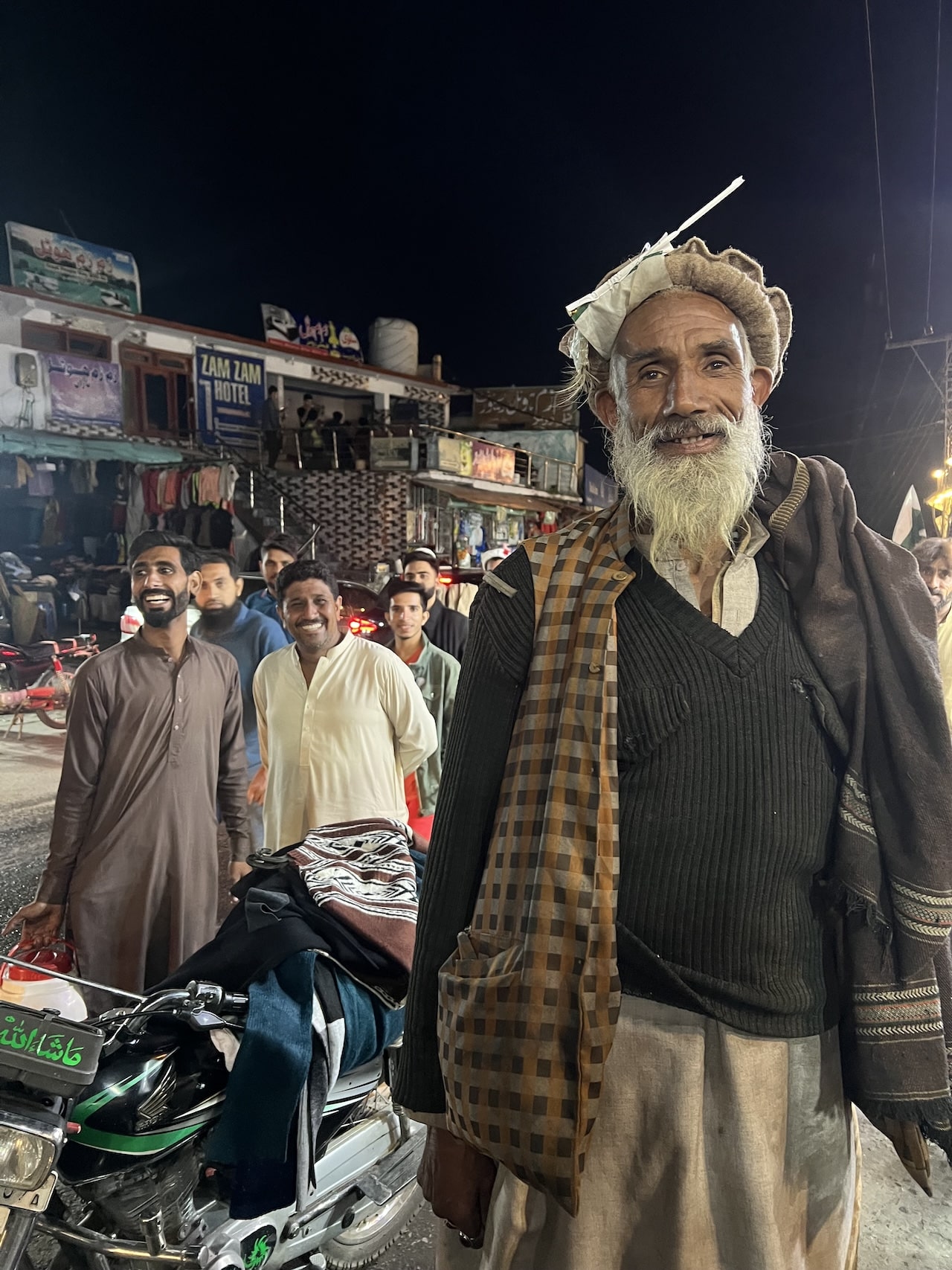 Uomo pakistano con cappello tradizionale Uomo anziano barbuto in cappello e gilet posa di notte per strada a Naran. Altri uomini sorridenti dietro di lui.