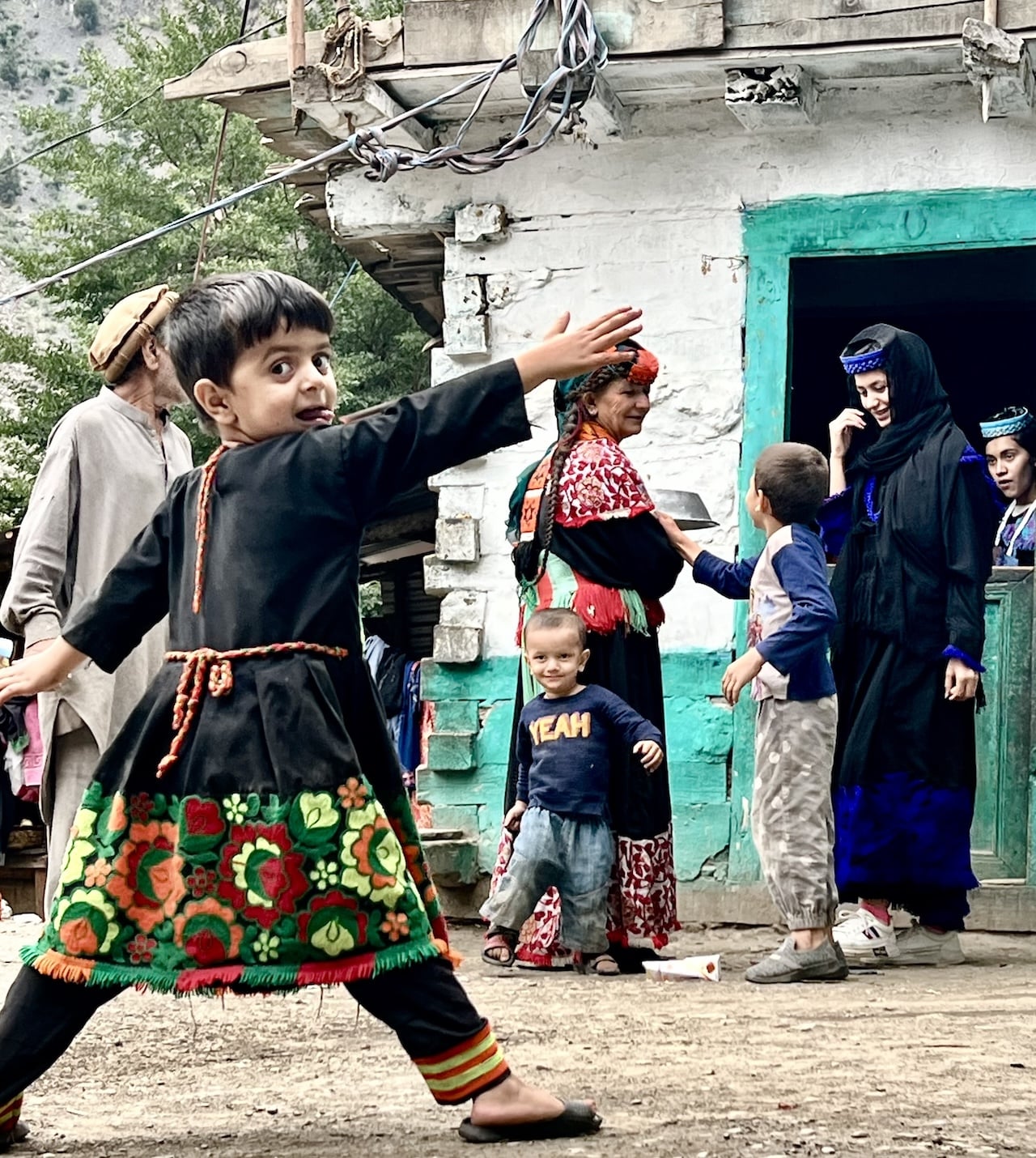 Bambino Kalash Bambini Kalash in abiti tradizionali decorati che giocano all'aperto davanti a una casa, adulti sullo sfondo