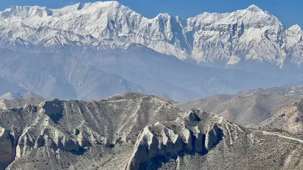 Catena montuosa con cime innevate e colline aride, formazioni rocciose in primo piano e cielo terso.
