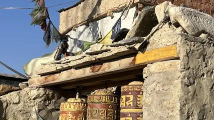 Ruote di preghiera tibetane dorate in fila, incorniciate da muro in pietra e bandiere di preghiera logore