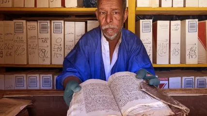Un bibliotecario mauritano di Chinguetti mostra un manoscritto antico, circondato da scaffali di libri.