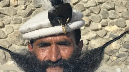 Primo piano di un uomo con baffi lunghi e un copricapo bianco decorato con piume, con uno sfondo di muro di pietra