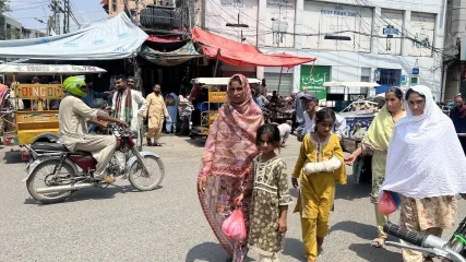 Donne e bambini camminano per una strada trafficata e assolata a Rawalpindi, con traffico e negozi sullo sfondo