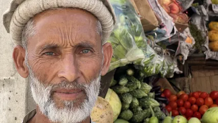 Uomo anziano con barba bianca e cappello Pakol in un bazar di Chitral, circondato da frutta e verdura fresche