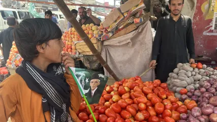 Bancarella di frutta e verdura, con un ragazzo che tiene una bandiera pakistana in mezzo a mucchi di pomodori e cipolle