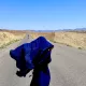 Donna su una strada nel deserto afghano guarda in lontananza dei veicoli, mentre il vento le scompiglia l'abito