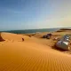 Tende bianche per accampamento turistico nel deserto, con dune di sabbia dorata, oceano sullo sfondo e cielo azzurro.