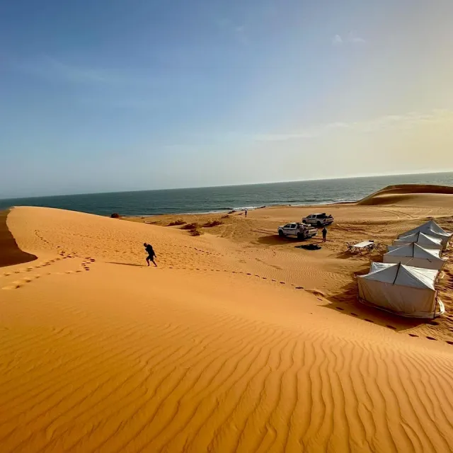 Tende bianche per accampamento turistico nel deserto, con dune di sabbia dorata, oceano sullo sfondo e cielo azzurro.